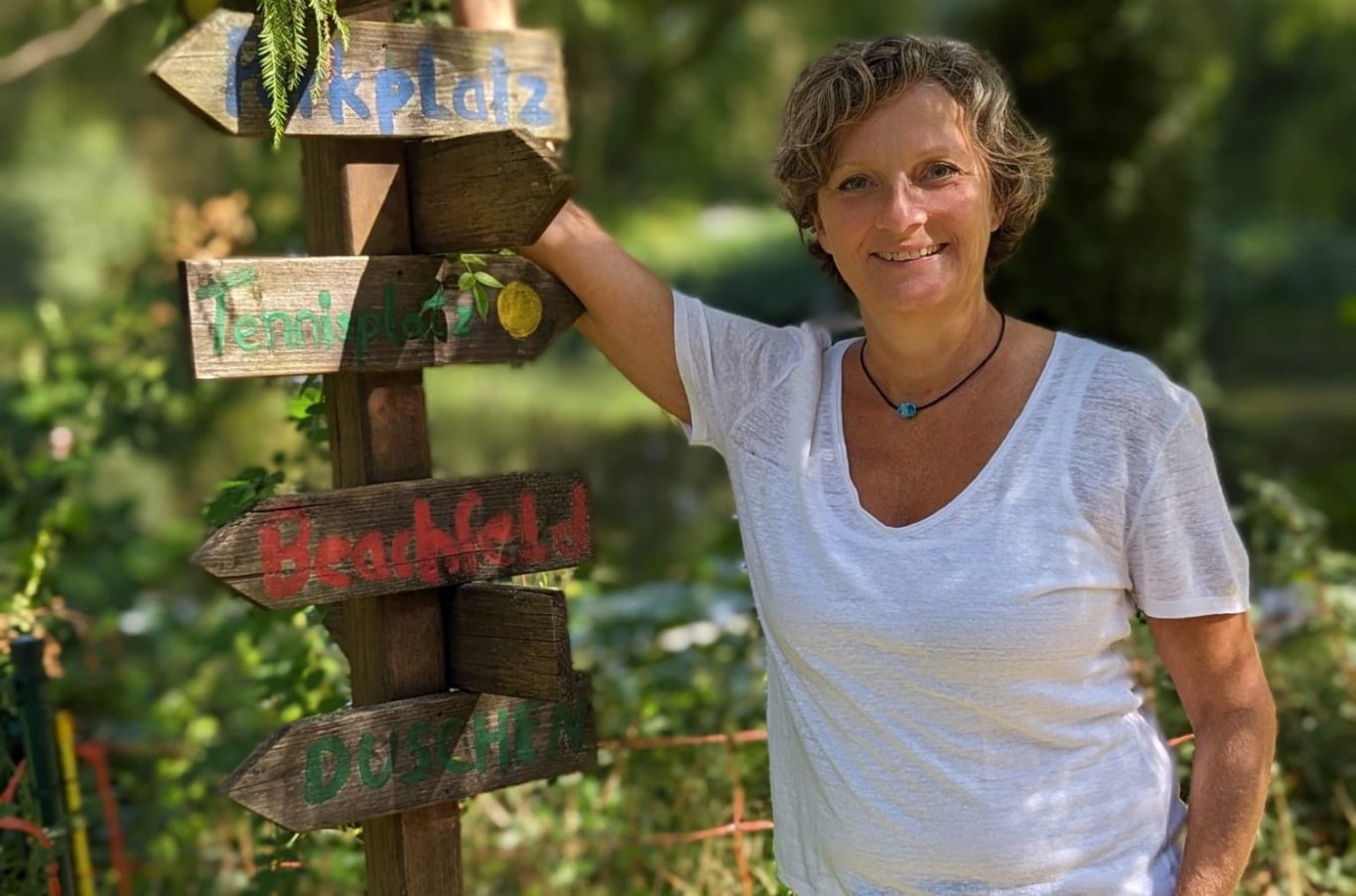 Andrea Mansfeld, gleich.wert.ich Eine Frau steht neben einem Schilderbaum mit Richtungshinweisen in einer grünen Umgebung.
